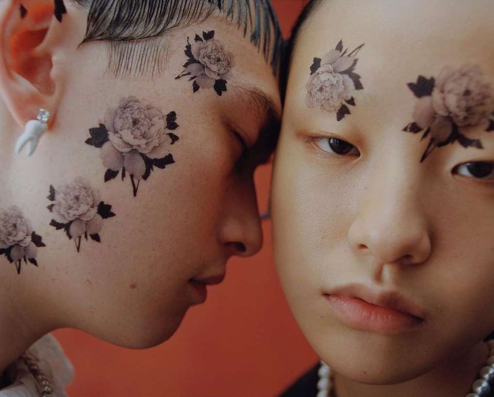 Two young adults, a male and a female, pose closely together in a studio setting with floral designs on their faces, close-up shot.