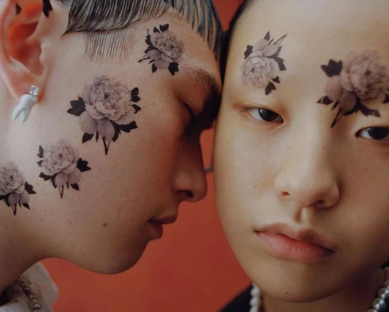 Two young adults, a male and a female, pose closely together in a studio setting with floral designs on their faces, close-up shot.