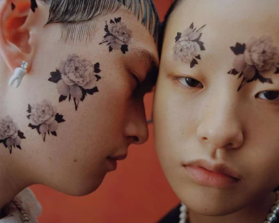 Two young adults, a male and a female, pose closely together in a studio setting with floral designs on their faces, close-up shot.