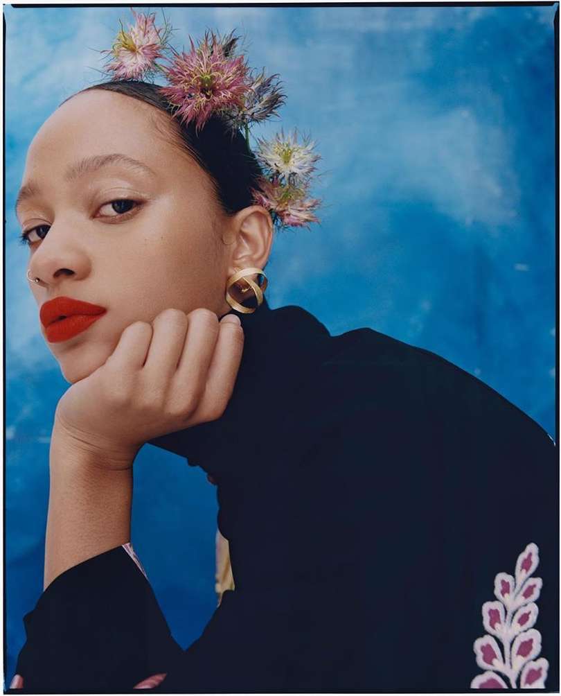 Portrait of a young woman wearing a black turtleneck with floral accents, featuring red lipstick, floral headpiece, and gold earrings.