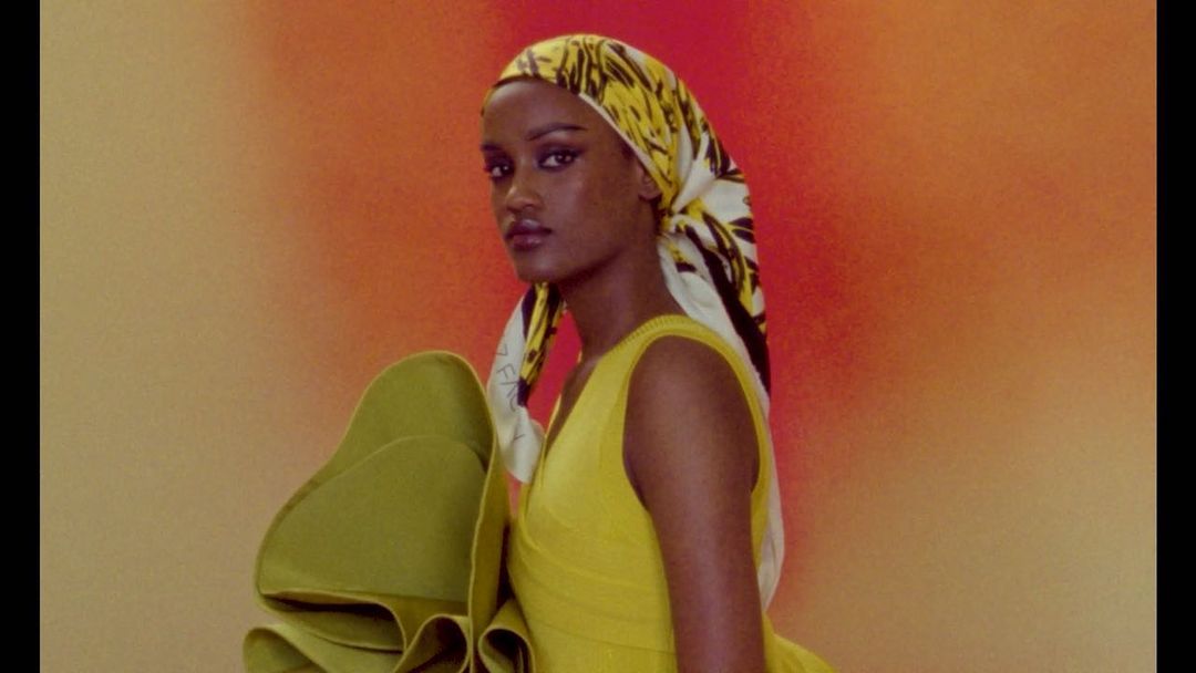 Portrait of an African-American woman in a yellow dress and patterned headscarf against an orange and beige background, looking at camera.