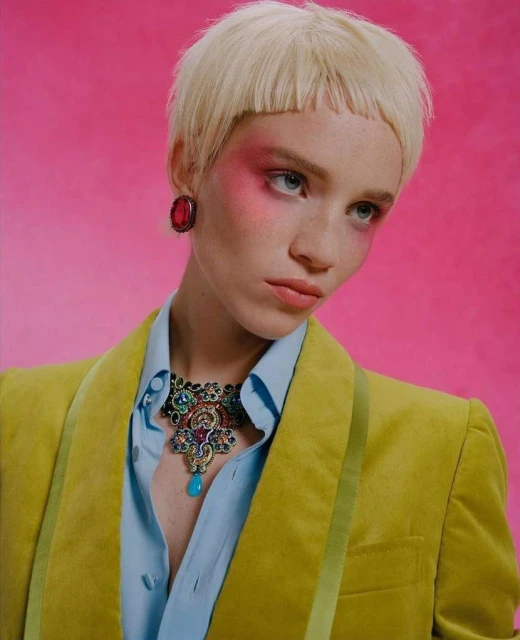 Close-up portrait of a woman with short blonde hair wearing a yellow jacket, blue shirt, and statement necklace against pink backdrop.