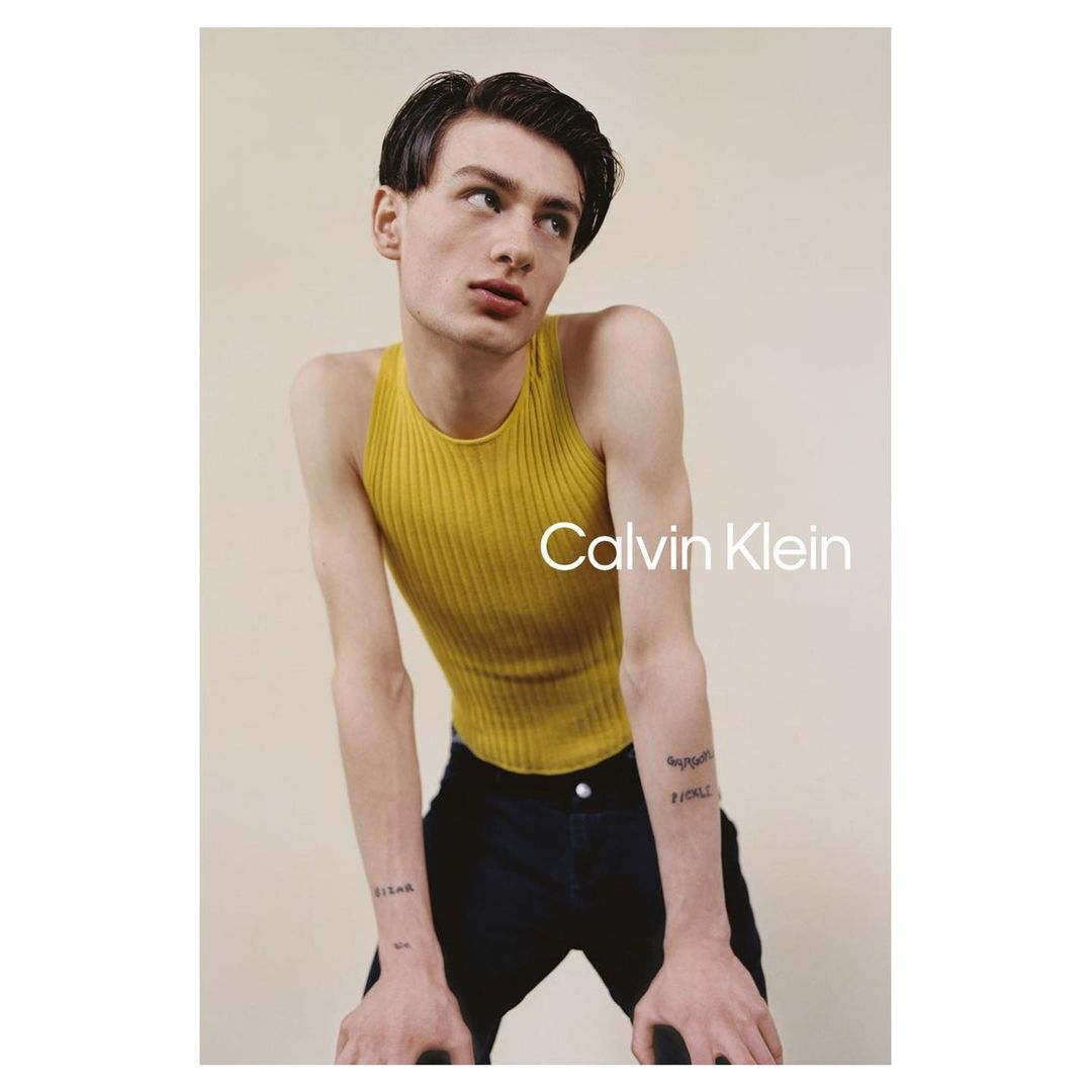 Young man in a mustard ribbed tank top leaning forward against a plain light background