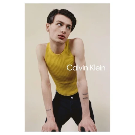 Young man in a mustard ribbed tank top leaning forward against a plain light background