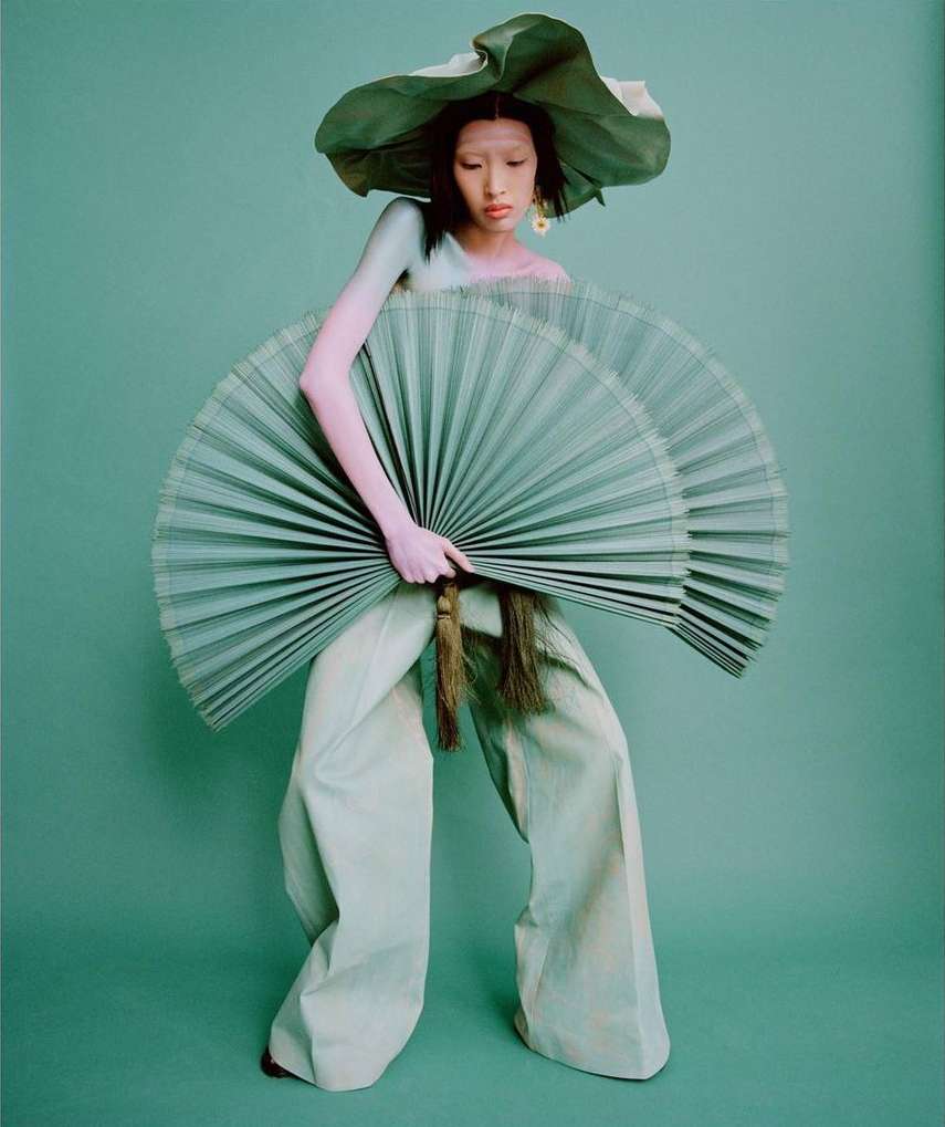 A person in wide leg pants holding two fans and wearing a large hat, all in shades of green, stands against a solid green backdrop.