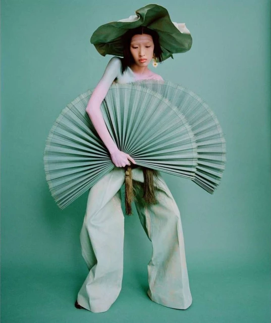 A person in wide leg pants holding two fans and wearing a large hat, all in shades of green, stands against a solid green backdrop.