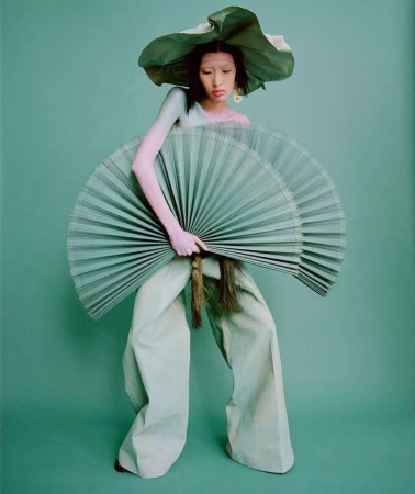 A person in wide leg pants holding two fans and wearing a large hat, all in shades of green, stands against a solid green backdrop.