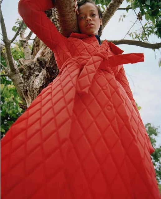 An eye-level shot features a woman in a red, diamond-patterned coat, leaning against a tree branch under soft, natural light.