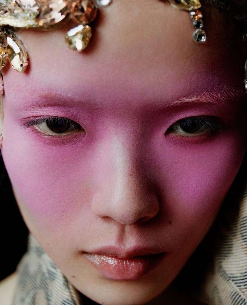 Close-up portrait of a young woman with pink face makeup and decorative headpiece, showcasing beauty and makeup artistry in a studio setting.