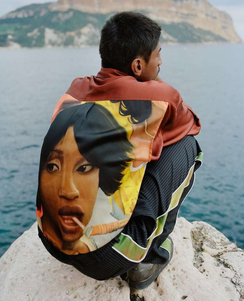Man in jacket with portrait art sits on rock with sea and mountains in background, portrait art of woman, seated human, outdoor setting.