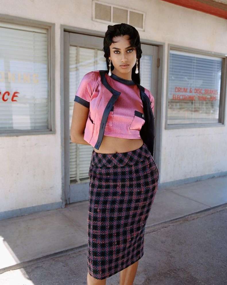 Young woman poses in pink top, dark jacket, and plaid skirt in front of a vintage building; fashion, style, and outdoor shot.