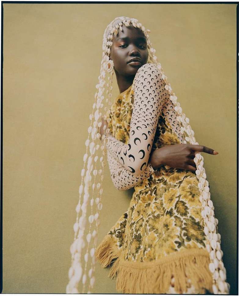 Fashion editorial shot of a dark-skinned woman with dramatic makeup wearing a patterned dress and headpiece with white beaded strands.