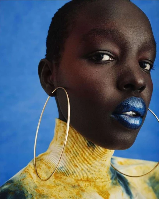 Close-up portrait of an adult woman with dark skin, blue metallic lipstick, gold hoop earrings, and wearing a tie-dye sweater on a blue background.