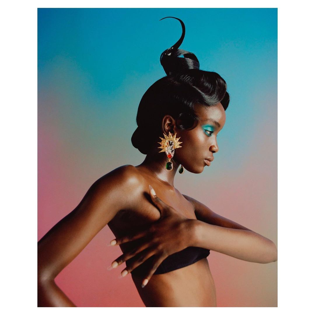 Profile portrait of a woman with dark skin wearing blue eyeshadow, elaborate hairstyle, decorative earrings, and black top against blurred backdrop.