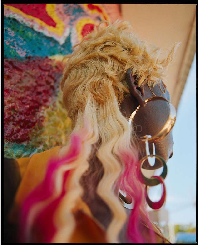 Back view of Black woman with blonde and pink-ombre hair, wearing gold hoop earrings, and a colorful painted wall in the background.