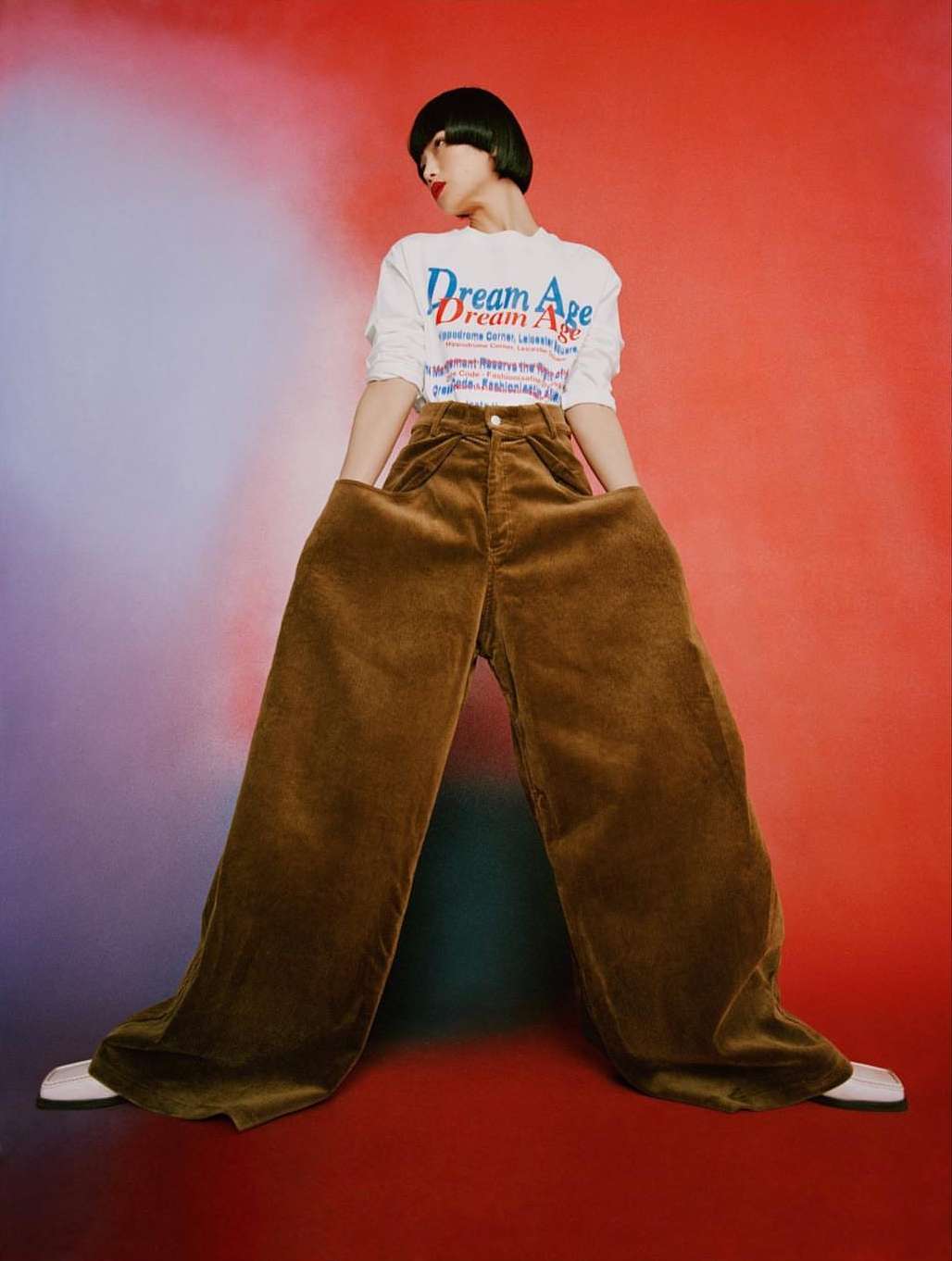 A person wearing oversized brown pants and a white graphic shirt in a studio with a gradient red and purple background.