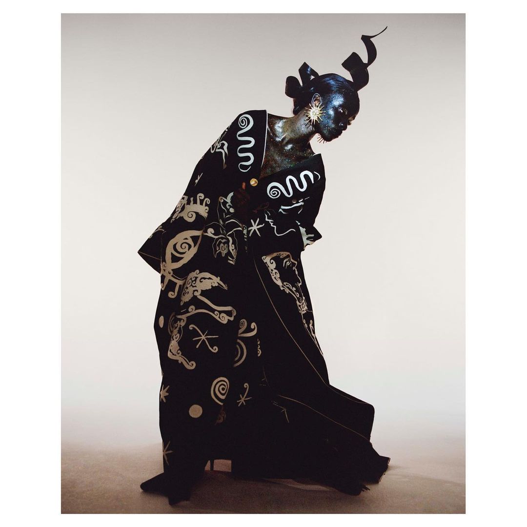 Fashionable African woman in patterned black robe and headpiece, adorned with gold accents and dramatic makeup, against a neutral backdrop.