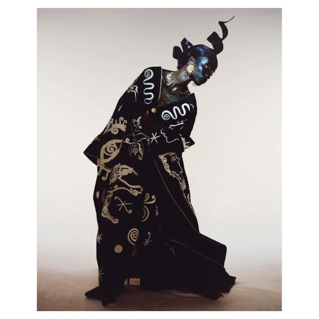 Fashionable African woman in patterned black robe and headpiece, adorned with gold accents and dramatic makeup, against a neutral backdrop.
