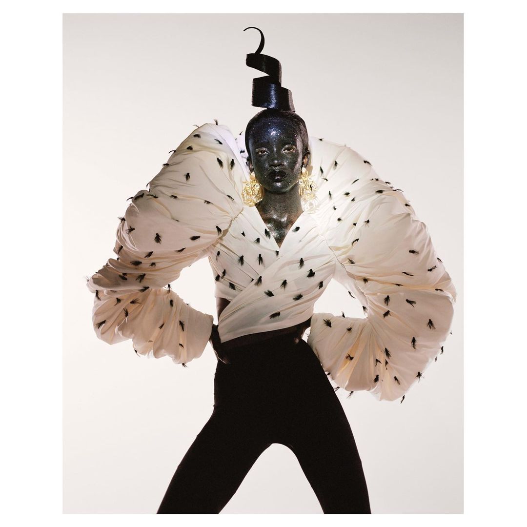 Fashion model with black skin and geometric headwear poses in a white, heavily ruffled top adorned with black insect-like patterns.
