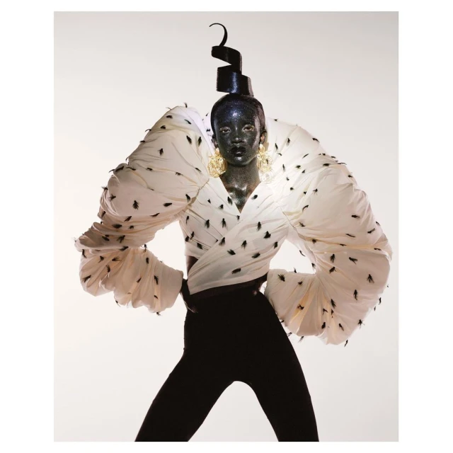 Fashion model with black skin and geometric headwear poses in a white, heavily ruffled top adorned with black insect-like patterns.