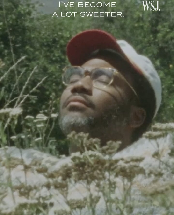 Adult male with closed eyes and gray beard, wearing a hat and glasses, surrounded by green plants and white flowers, outdoors.