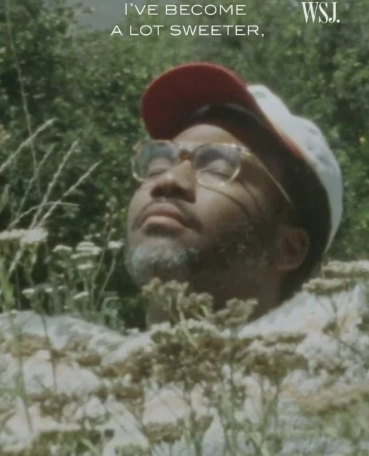 Adult male with closed eyes and gray beard, wearing a hat and glasses, surrounded by green plants and white flowers, outdoors.