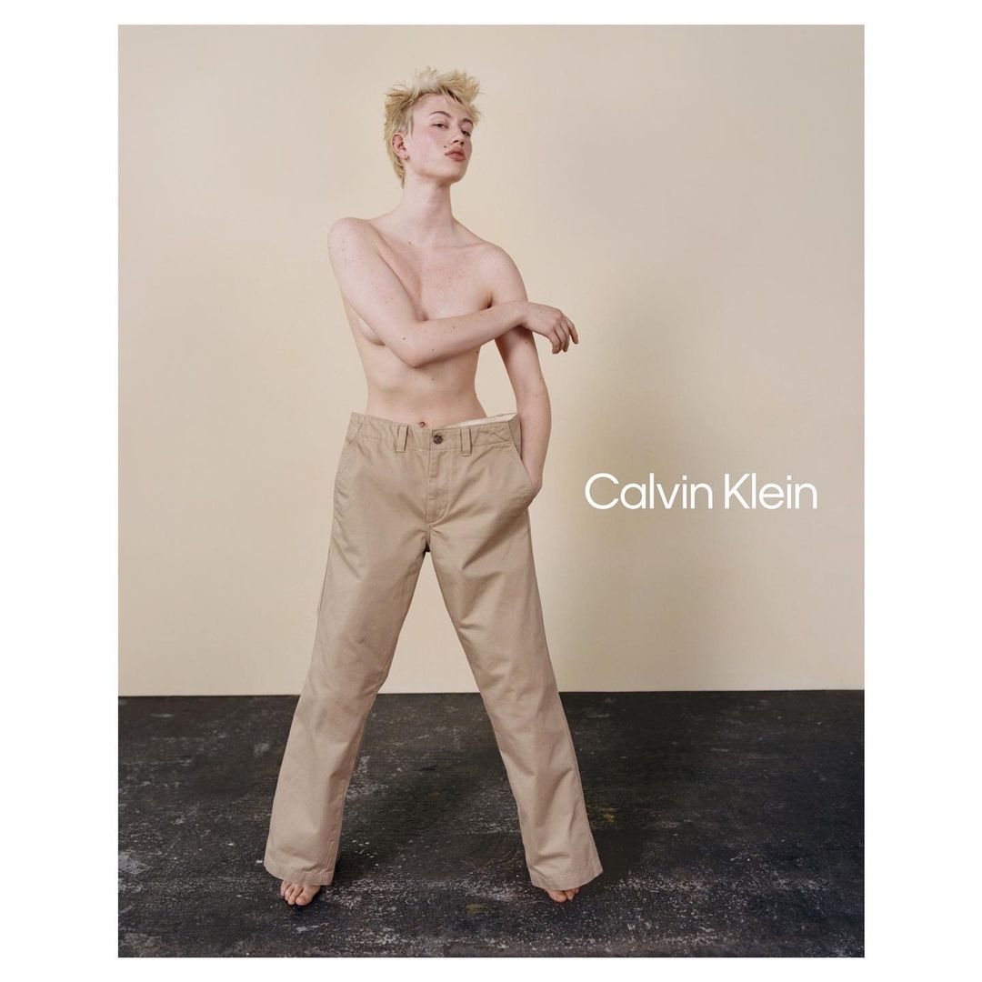 A gender-neutral person posing in a Calvin Klein advertisement, wearing beige pants with visible logo, against a neutral backdrop in a studio.