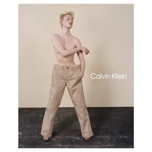 A gender-neutral person posing in a Calvin Klein advertisement, wearing beige pants with visible logo, against a neutral backdrop in a studio.