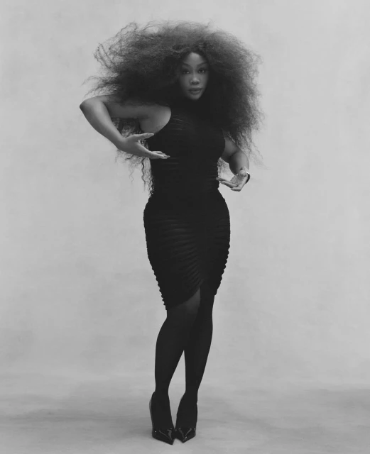 Black and white full shot of an African American woman in a black dress with voluminous hair striking a dance pose in a studio.