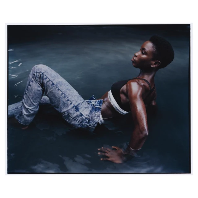 Adult female model poses in water wearing a black Calvin Klein sports bra and acid wash jeans; studio shot, fashion, beauty.