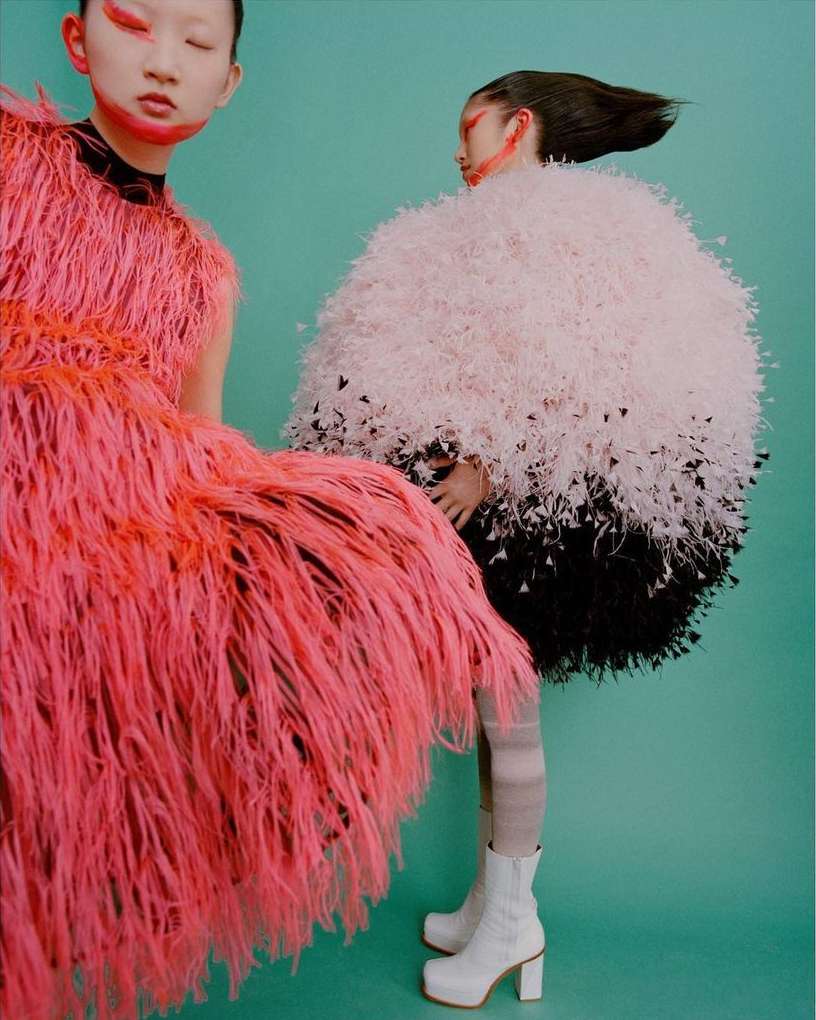 Two models wearing textured feathered dresses and bold face makeup against a solid turquoise background.