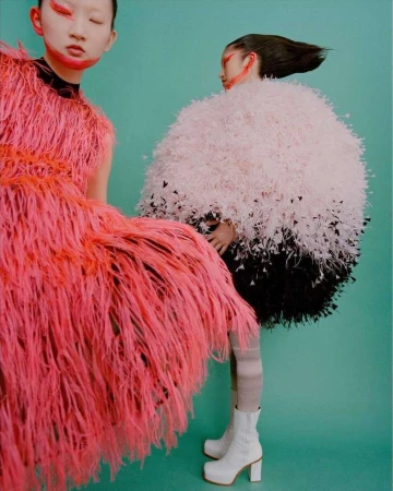 Two models wearing textured feathered dresses and bold face makeup against a solid turquoise background.