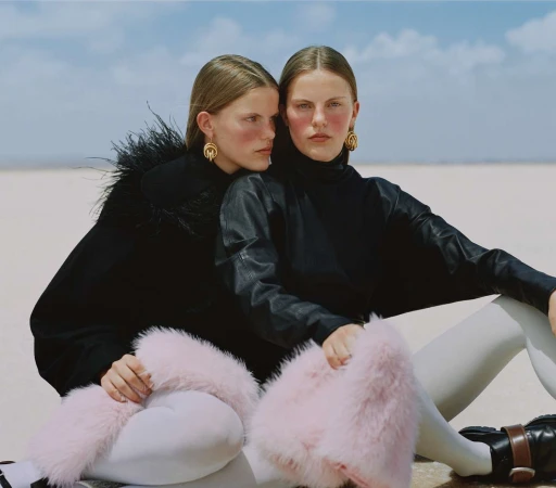 Two female models in black outfits with pink fluffy cuffs seated outdoors against a pale horizon under a bright sky.