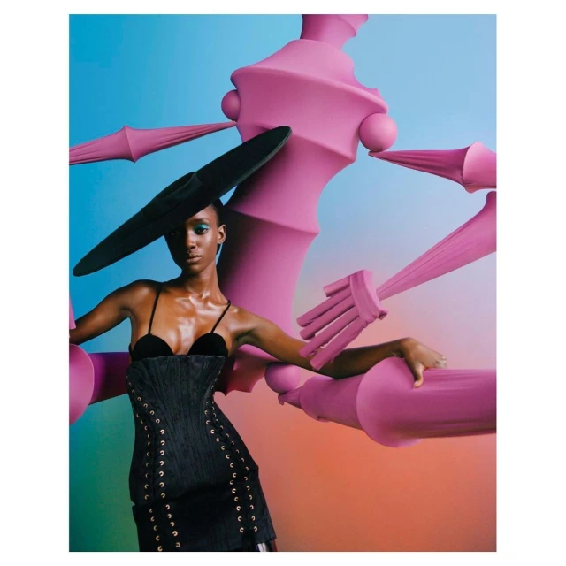 A black female model poses in a black corset dress and large black hat against a gradient background with an abstract, pink structural object.