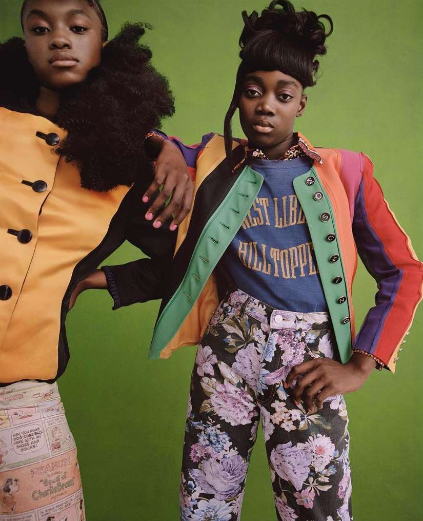 Two Black women pose in colorful fashion against a green backdrop, one with a black turtleneck and the other in a floral-print jacket.