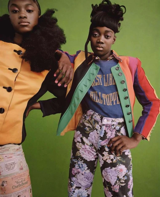 Two Black women pose in colorful fashion against a green backdrop, one with a black turtleneck and the other in a floral-print jacket.