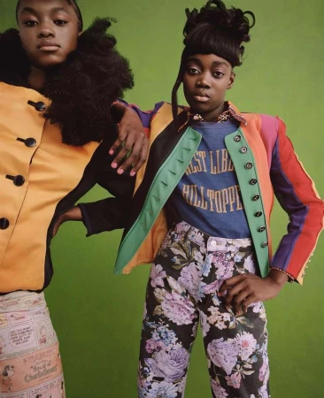 Two Black women pose in colorful fashion against a green backdrop, one with a black turtleneck and the other in a floral-print jacket.