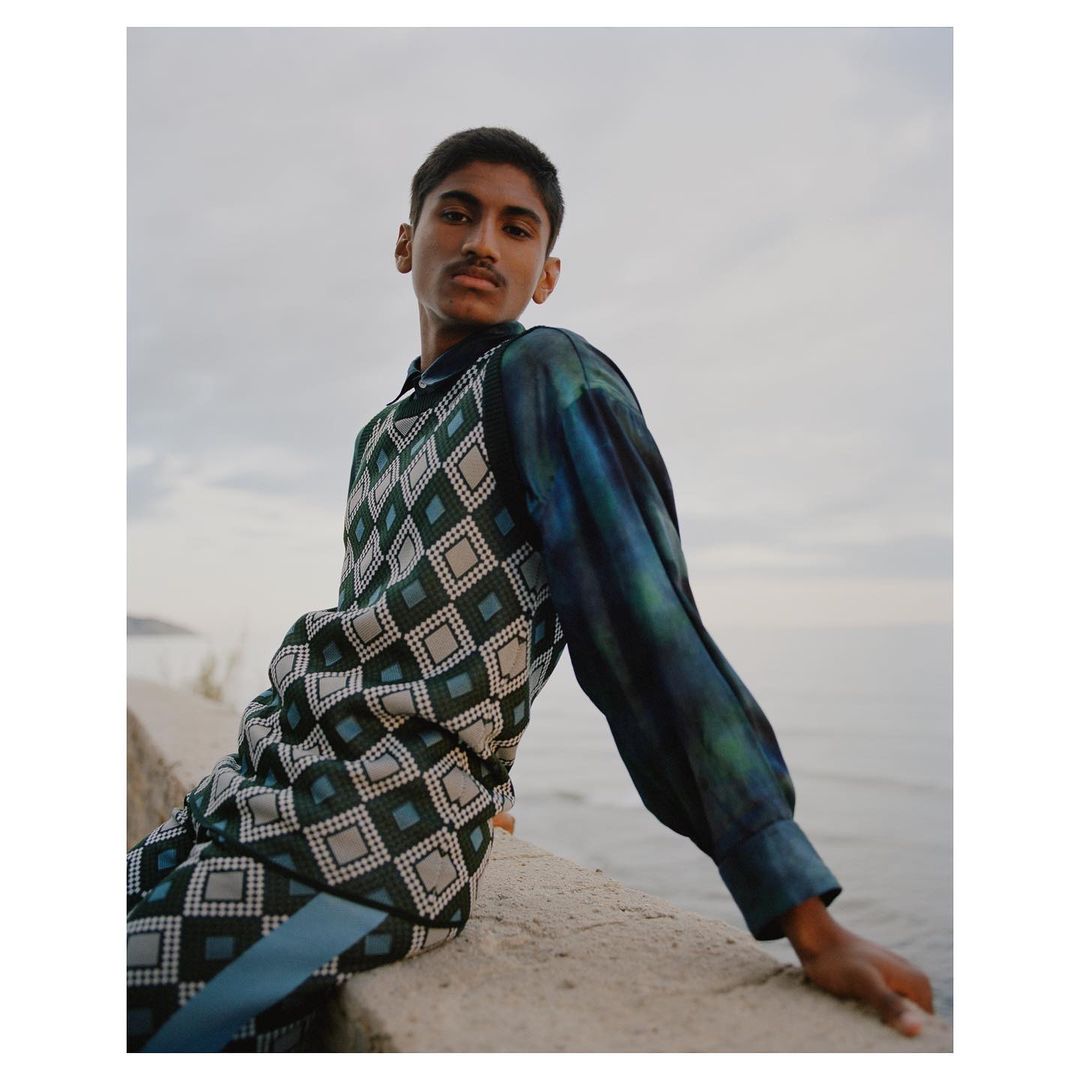 Portrait of a young man in patterned top and pants sitting on a stone ledge with a sea and cloudy sky backdrop in natural light.