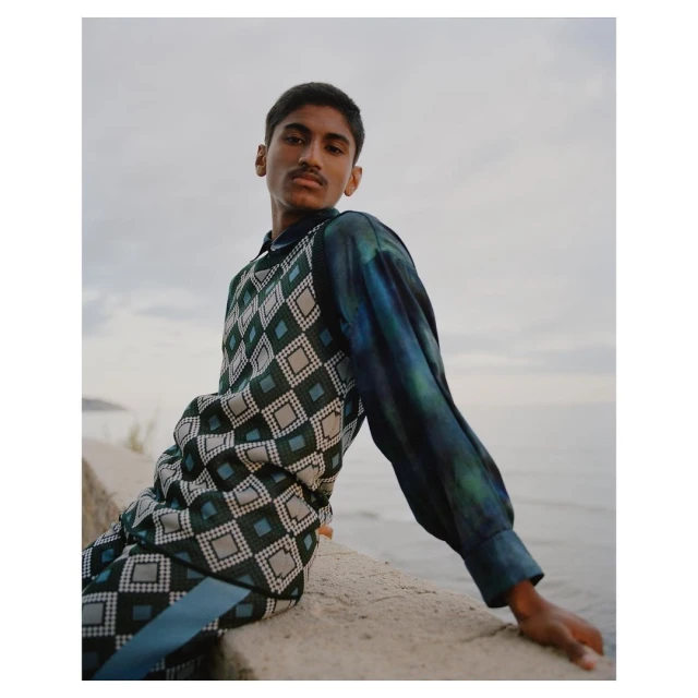 Portrait of a young man in patterned top and pants sitting on a stone ledge with a sea and cloudy sky backdrop in natural light.