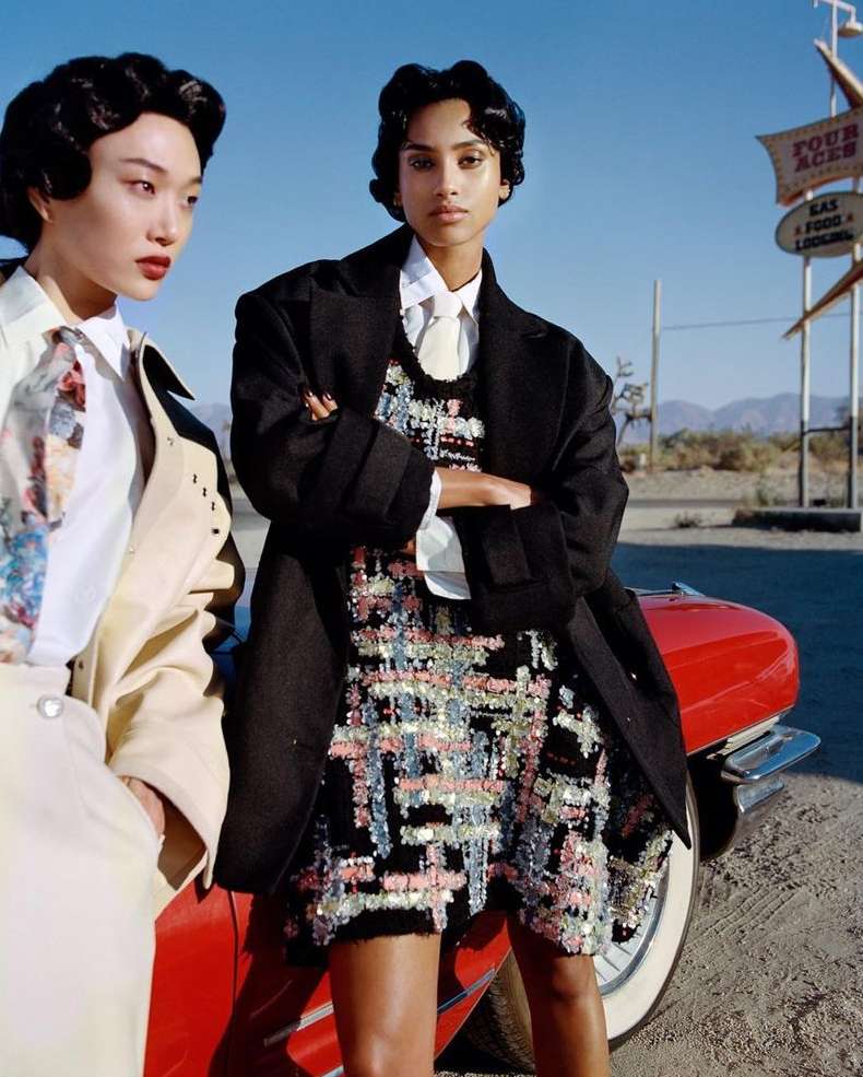 Two fashion models pose in front of a red classic car and a vintage motel sign under a clear sky in a desert setting.