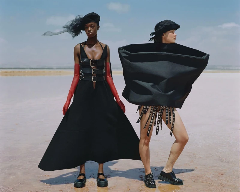 Two fashion models pose outdoors, one in a black dress with red gloves, the other in an oversized black top and studded shorts.