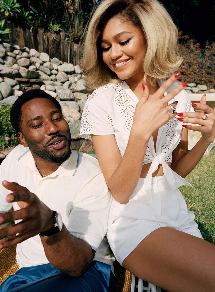 Zendaya and John David Washington pose outdoors on a sunny day. Zendaya wears a white top and shorts; Washington wears a white polo shirt.