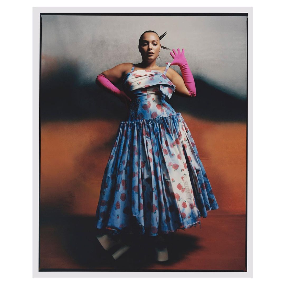 Adult female fashion model wearing a pastel floral-print dress and pink gloves posing in a studio against a blended gradient background.