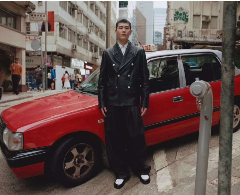 Adult male in a black jacket and wide pants stands on an urban street in front of a red taxi in a daytime medium shot.