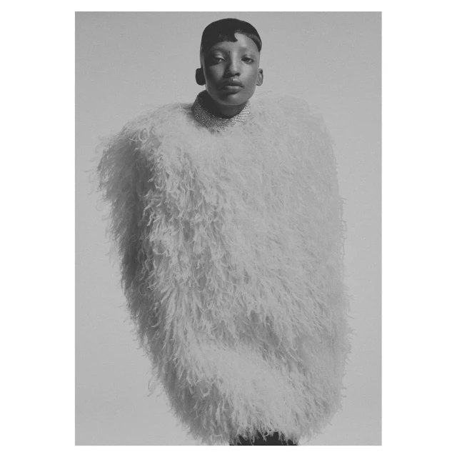 Elegant woman posing indoors wearing an oversized, fluffy white coat with a round neckline and a sparkling choker necklace.