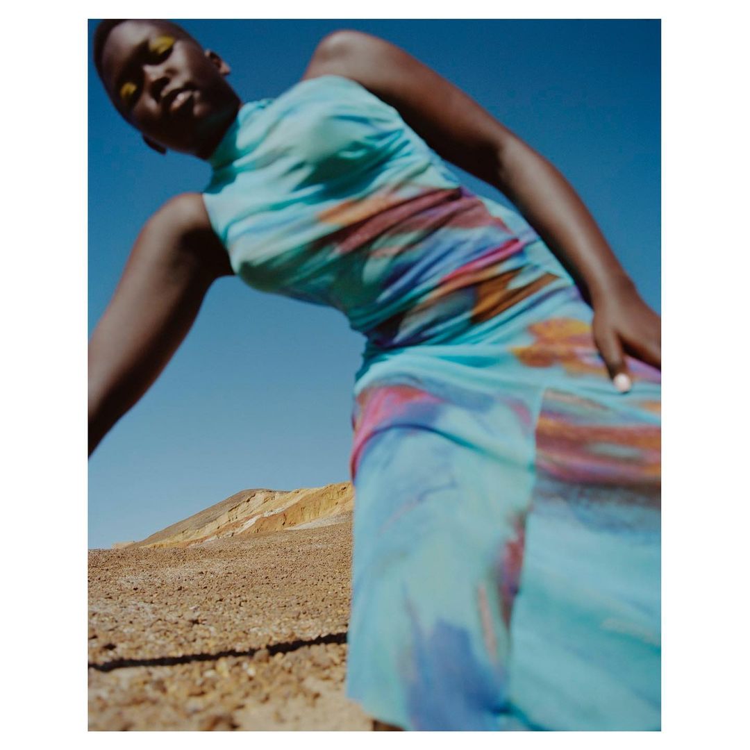 Woman in flowing dress posing in desert setting. Fashion photography shows full-length portrait against blue sky and arid background.