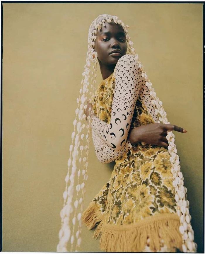 Portrait of a Black woman wearing a printed dress and a long headdress strung with cowrie shells