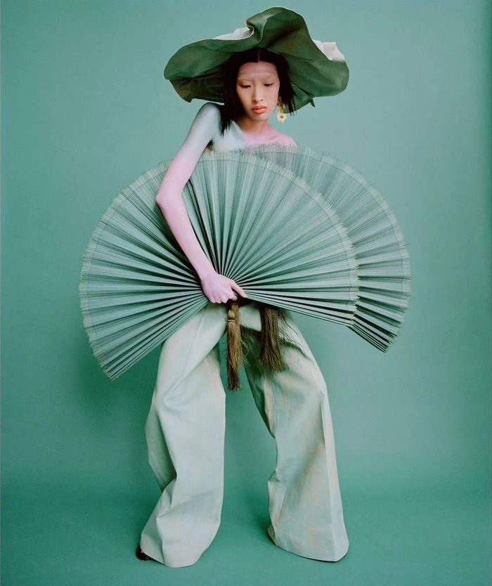 Model in wide-brimmed green hat holding large pleated fan wearing wide trousers against a solid green background