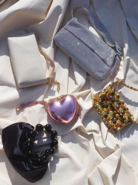 Variety of designer evening bags: a white rectangular clutch, a grey sparkly purse, a pink heart bag, a beaded bag, and a black satin purse.