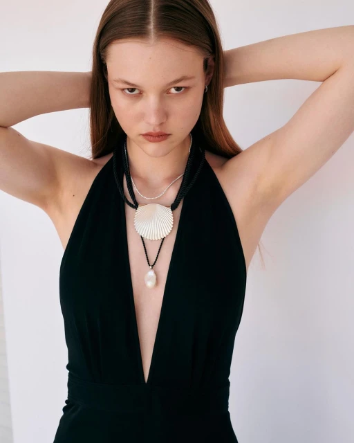 Portrait of a young woman with hands behind her head, wearing a low cut black dress and statement necklaces in a white studio.